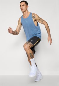 Blå tanktop med sideskæringer, sorte shorts med gule accenter og hvide sportssko. Personen er i en løbeholdning.
