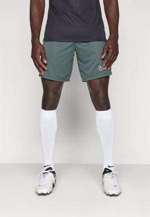 Homme portant un short de sport Nike vert foncé, des chaussettes blanches montantes jusqu'aux genoux et des crampons blancs avec des détails noirs, debout les mains détendues.