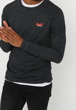 Homme portant une chemise à manches longues gris foncé avec un petit logo rouge et blanc sur la poitrine, un t-shirt blanc en dessous et un pantalon noir, mains jointes devant.