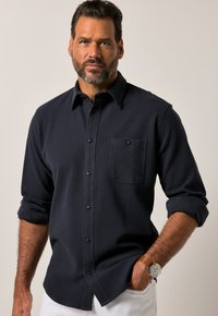 Granatowa koszula z guzikiem, o teksturowanym wykończeniu dzianinowym, z jedną kieszenią na piersi i podwiniętymi rękawami. Posiada czarne guziki i krągły krój dołu.