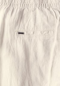 Ljusbeige tyg med en rynkig textur, försedd med ett elastiskt midjeband och en liten, silvrig logotagg sydd på ytan.