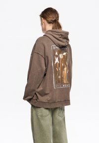 Sudadera con capucha marrón que presenta un gran diseño gráfico en la parte posterior. El diseño incluye flores en colores suaves sobre un fondo texturizado.