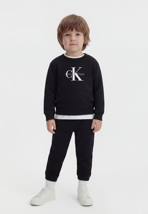 Bambino piccolo con capelli castano chiaro in piedi, indossa una felpa nera Calvin Klein, pantaloni neri, calzini bianchi e scarpe da ginnastica bianche su sfondo bianco.