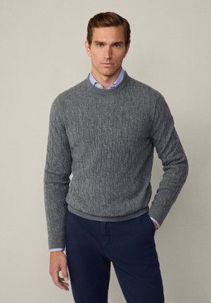 Pull en tricot gris avec un motif en câble, col rond et poignets côtelés. Porté par-dessus une chemise à col bleu clair et associé à un pantalon bleu marine.