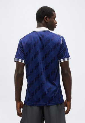 Egy férfi háttal, sötétkék, rövid ujjú pólót visel, finom átlós mintával, valamint fehér csíkos gallérral és ujjvégel.