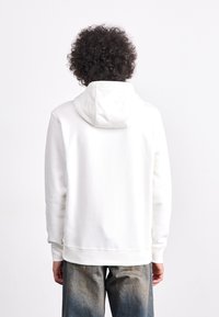 Weißer Kapuzenpullover mit Kordelzug und weicher Textur, ausgestattet mit einer Kängurutasche, getragen mit ausgebleichten blauen Jeans im straighten Schnitt.