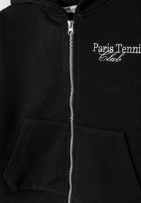 Crna dukserica sa zipom napravljena od teksturirane tkanine. Ima prednji džep i izvezeni logo "Paris Tennis Club" u bijeloj boji.
