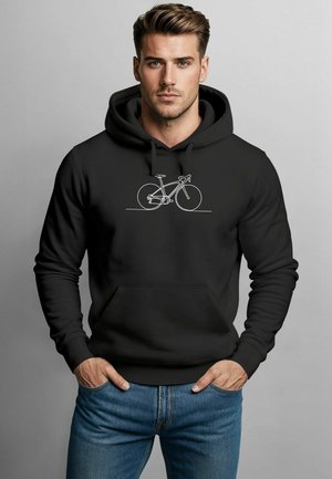Mann in schwarzem Kapuzenpulli mit weißem Linienz الرسم von einem Fahrrad auf der Brust, Hände in den Taschen, stehend vor einem schlichten grauen Hintergrund.