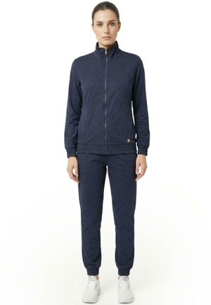 Une femme porte un survêtement bleu marine zippé, avec un pantalon de jogging assorti et des baskets blanches, sur un fond blanc uni.