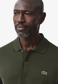 Mörkgrön polo t-shirt med strukturerad tyg, två knappar vid kragen och en vitkantad krokodil-logotyp placerad på vänster bröst.