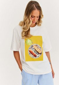 Bílý tričko s barevným potiskem jídla, na kterém je talíř s chlebem a zelený nápoj na žlutém pozadí. Pruhované kalhoty.