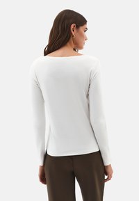 Kvinde med langt brunt hår iført en hvid bluse med lange ærmer og brune bukser, set bagfra mod en ensfarvet baggrund.