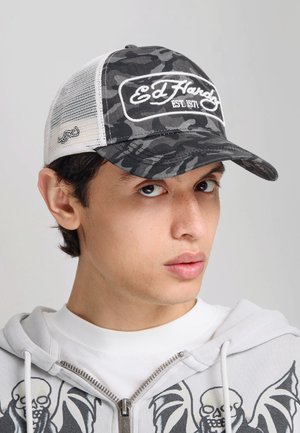 Joven con una gorra Ed Hardy de camuflaje negra y gris y una sudadera blanca con cremallera con diseños de calaveras negras sobre un fondo gris.