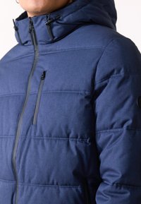 Chaqueta acolchada de color navy con cremallera frontal, capucha ajustable, paneles cosidos y bolsillos laterales. El material tiene un acabado suave y mate.