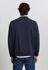 Sudadera azul marino con cuello redondo, puños y dobladillo acanalados; tela suave; se lleva sobre una camisa blanca, combinada con vaqueros azules con detalles en amarillo.