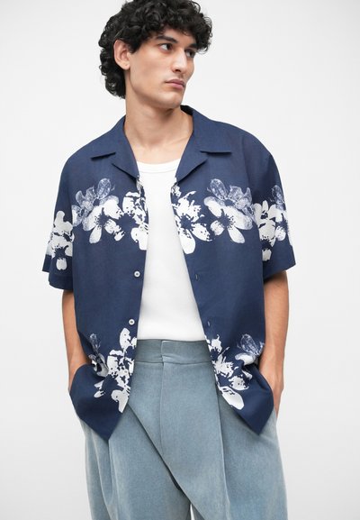 Un jeune homme aux cheveux bouclés et foncés porte une chemise à manches courtes bleu marine avec un imprimé floral blanc par-dessus un débardeur blanc et un pantalon bleu clair.