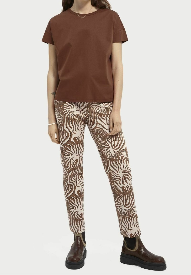 Femme portant un haut marron uni à manches courtes, un pantalon marron et blanc à motif abstrait, et des bottines marron foncé.
