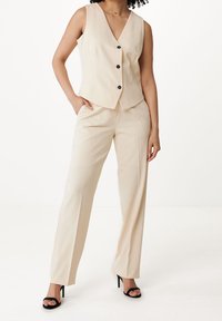 Beige vesta s V výstřihem a třemi černými knoflíky, ve spojení s béžovými, dobře střiženými kalhotami a černými sandály na podpatku. Hladká textura, padnoucí design.