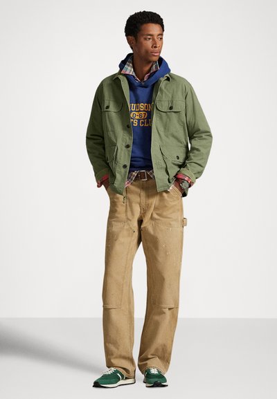 Μπουφάν ελιάς πάνω από μπλε σκουρόχρωμο φούτερ με κίτρινο κείμενο, πουκάμισο καρό, χακί παντελόνι και πράσινα αθλητικά παπούτσια με λευκές λεπτομέρειες. Μινιμαλιστικός σχεδιασμός.