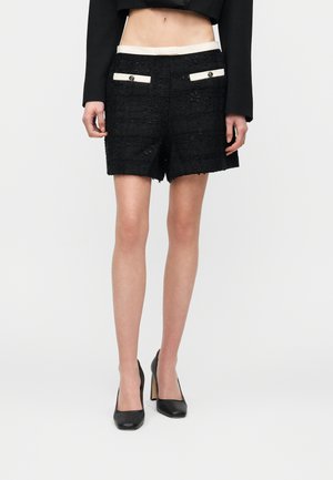 Frau trägt schwarze, strukturierte Shorts mit weiß eingefassten, mit Knöpfen versehenen Taschen, kombiniert mit schwarzen High Heels und einem kurzen schwarzen Oberteil.