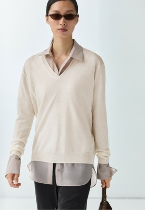 Donna con occhiali da sole scuri, maglione beige a scollo a V sopra camicia taupe, pantaloni neri, che tiene una borsa marrone su sfondo chiaro.