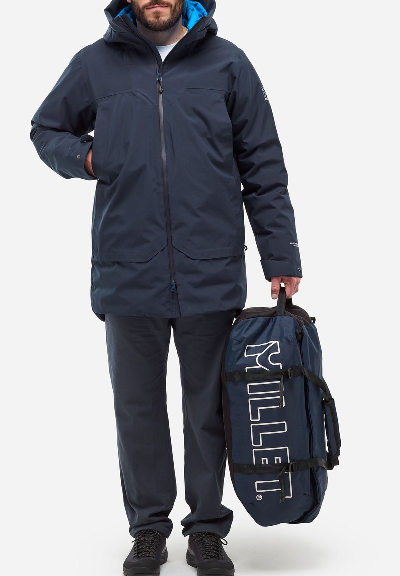 Marineblau wasserfeste Jacke mit Kapuze, ausgestattet mit einem blauen Innenfutter, Reißverschluss vorne und zwei Fronttaschen. Dunkle Hose und eine marineblaue Reisetasche werden gehalten.