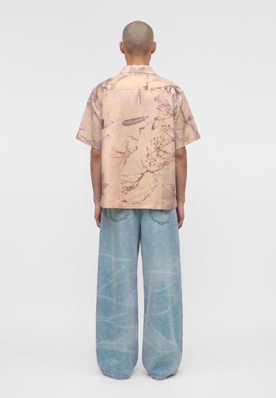 Άτομο με ξυρισμένο κεφάλι, που φοράει μπεζ κοντομάνικο πουκάμισο με φυτικό μοτίβο και ανοιχτό μπλε τζιν με φαρδιές φάσες, στέκεται γυρισμένο με την πλάτη σε απλό φόντο.