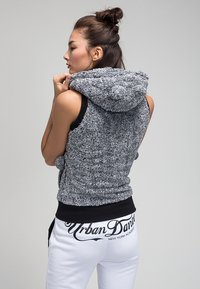 Pelēka teksturēta bez piedurknēm, ar biezu kapuci, kopā ar baltām treniņbiksēm, kurām aizmugurē ir melns uzraksts "Urban Dance" jostasvietā.