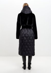 Zwarte nepbont cropped jack met een capuchon, gecombineerd met een lange zwarte gewatteerde rok met een diamanten patroon en een slanke riem.