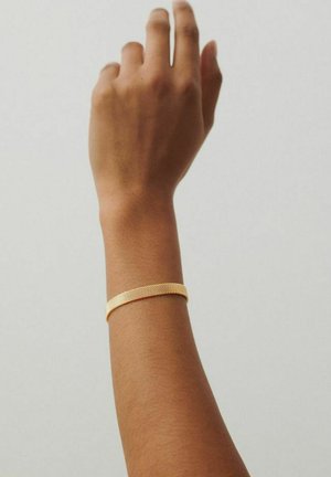 Ranką pakėlusi prieš neutralią foną, ant riešo dėvint tekstūruotą auksinį apyrankę.