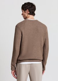 Maglione marrone lavorato a maglia con superficie testurizzata, scollo rotondo e polsini a coste. Indossato sopra una camicia bianca, abbinato a pantaloni chiari.