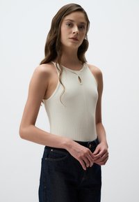 Body in crema a costine con colletto alto e dettaglio a chiave frontale, abbinato a jeans denim blu scuro.