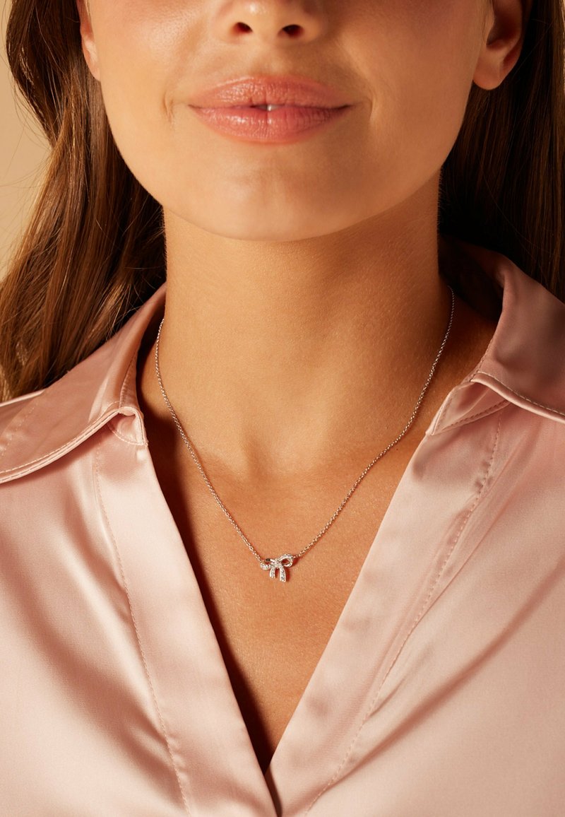 Zilveren halsketting met een klein strikbedel met complexe details, gedragen boven een lichtroze satijnen blouse.