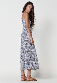 Rochie maxi din țesătură florală albastră și albă, cu bretele ajustabile, corset ajustat și fustă în straturi. Asortată cu sandale maro cu curelușe.