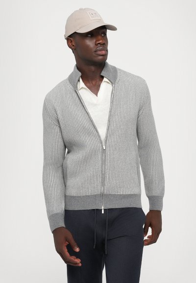 Grau gestreifter Reißverschluss-Pullover mit hohem Kragen und silbernem Reißverschluss. Kombiniert mit einem hellen Hemd und marineblauen Hosen. Eine lässige Mütze sorgt für Stil.