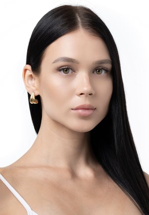 Goldene Creolen mit einem skulpturalen, gedrehten Design. Glatte Oberfläche und glänzende Ausführung, vor einem neutralen Hintergrund.