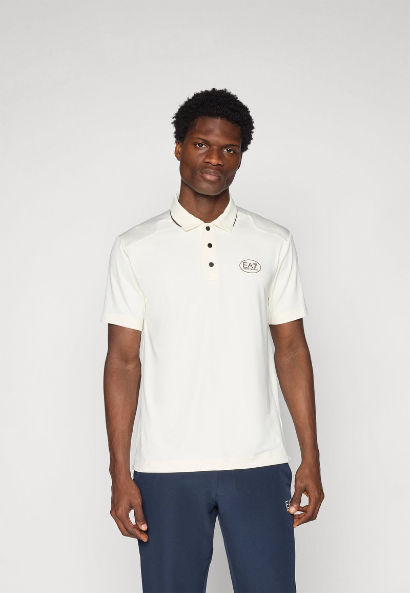 Πόλο μπλουζάκι σε ανοιχτό κρέμα με γιακά και κουμπιά, που διαθέτει σκούρο κεντημένο λογότυπο στην αριστερή πλευρά του στήθους και κοντά μανίκια.