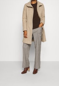 Abrigo de lana beige con un cuello alto, suéter de punto marrón, pantalones de pata ancha de cuadros y botines con tacón marrón y puntera cuadrada.