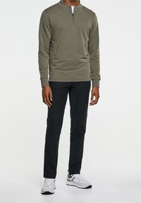 Suéter de manga larga color verde oliva con cuello de media cremallera, hecho de una mezcla de algodón. Combinado con pantalones ajustados negros y zapatillas blancas.