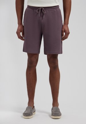 Man draagt paarse wafelkatoenen short met trekkoord en grijze slip-on schoenen, staand tegen een effen achtergrond.