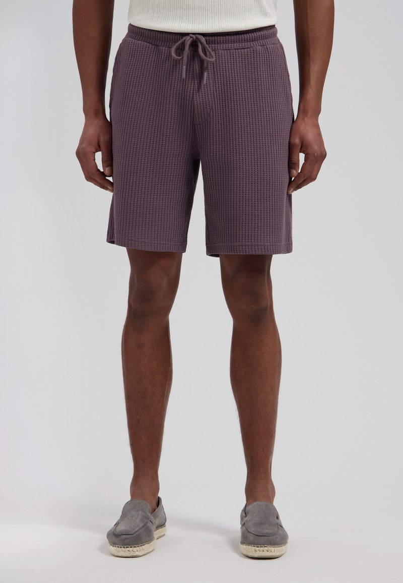 Homme portant un short violet en maille gaufrée avec cordon et des chaussures grises slip-on, debout devant un fond uni.