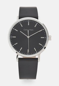 Montre Calvin Klein avec un cadran rond noir, des index et des aiguilles argentés. Présente un bracelet en cuir noir lisse et des accents en acier inoxydable.