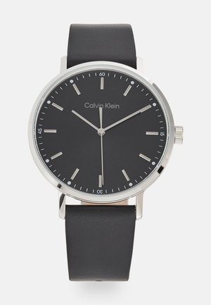 Calvin Klein watch with a round black dial, silver markers, and hands. Features a smooth black leather strap and stainless steel accents.