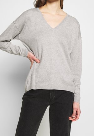 Persona con un suéter gris claro de cuello en V y mangas largas, combinado con pantalones negros, con la mano apoyada en la cadera.