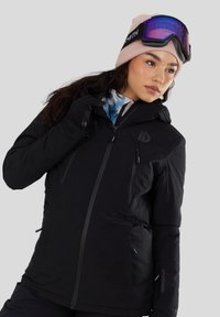 Jonge vrouw die een zwarte winterjas, zwarte handschoenen, een roze muts en skibril op het voorhoofd draagt, kijkt zijwaarts.