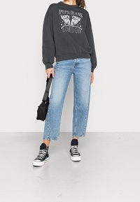 Felpa grigio scuro con ali stampate e scritte, jeans dritti blu chiaro e sneaker alte nere con suola bianca.