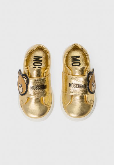 Kuldsetest metallikslikest tennisetelt Velcro rihmaga, millel on karumõmmilogo ja brändinimi "Moschino" trükituna. Kummised tallad, ümarad varbad.