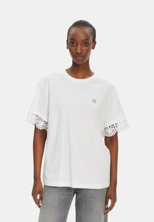 T-shirt bianco in cotone con rifinitura in pizzo sulle maniche corte e un piccolo logo sul davanti. Vestibilità casual con scollatura rotonda.