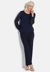 Marineblaues Oberteil mit strukturierten, durchsichtigen Ärmeln, kombiniert mit passenden marineblauen Hosen. Das Model trägt silberne, schicke Sandalen. Einfaches, elegantes Design.