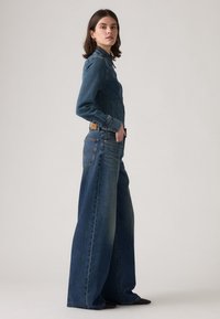 Combinaison en denim bleu moyen, avec un haut ajusté, un design à jambes larges et des poches à rabat. La texture est lisse avec un léger effet délavé.